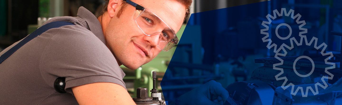 Maschinenbau Header: Ein Maschinenbauer ist mit einer Schutzbrille in die Kamera lächelnd über eine Werkbank gebeugt.